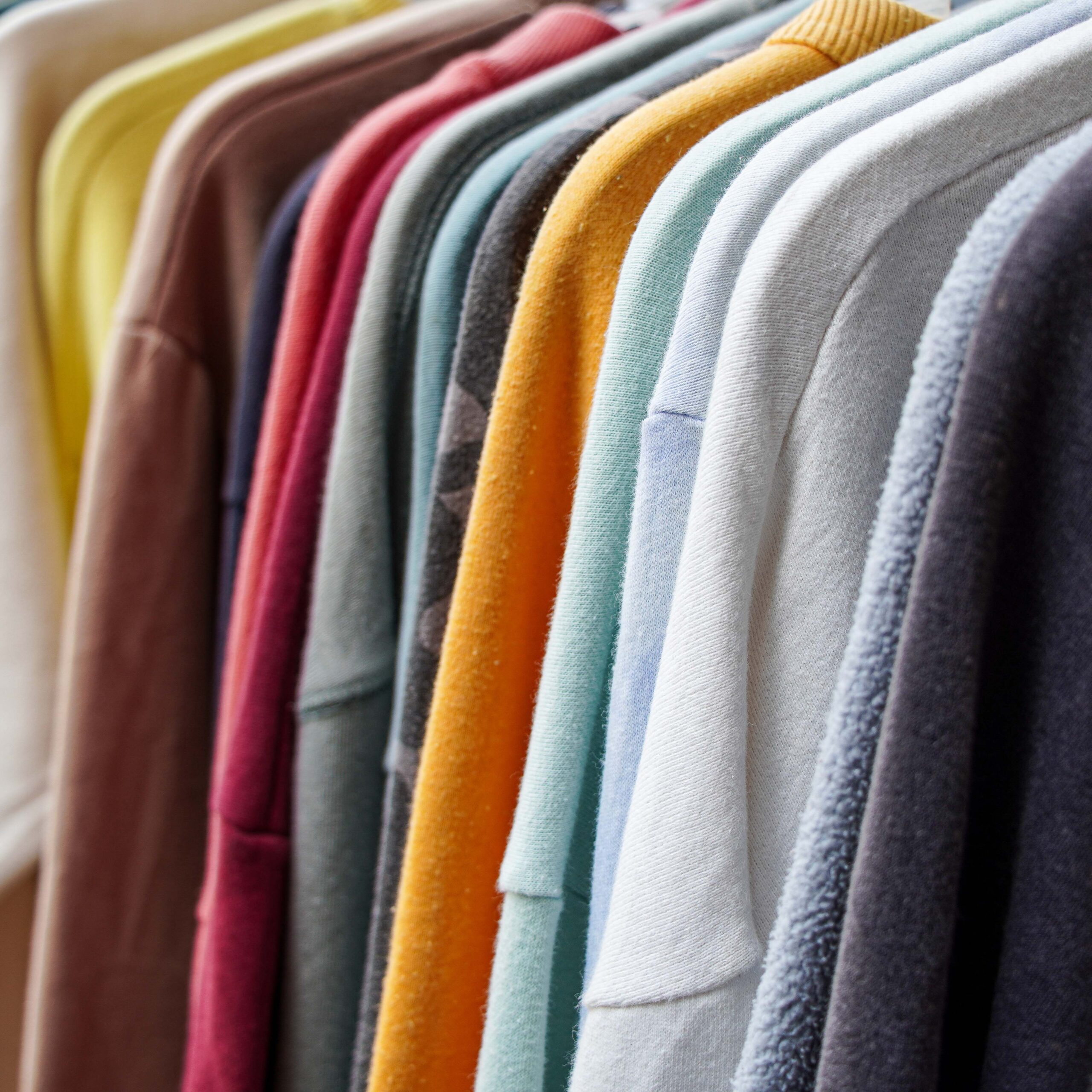 A close up view of a row of colorful sweatshirts hanging on a rack in a store. The vibrant colors of the fabrics create a pleasing visual contrast. The clothes are showcasing their soft textures.